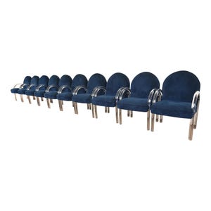 May include: A set of ten navy blue armchairs. Each chair has a rounded back, clear acrylic armrests, and silver metal legs. The chairs are arranged in a row, perfect for a dining room or living room.
