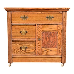 May include: A wooden cabinet with a warm, reddish-brown finish. It features two drawers, two smaller drawers, and a cabinet door with a patterned panel. The cabinet has ornate brass handles and sits on small caster wheels.