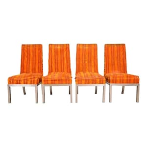 May include: Four dining chairs with orange and red striped upholstery. The chairs have a rectangular seat and back, with a silver metal frame. The chairs are arranged in a row against a white background.