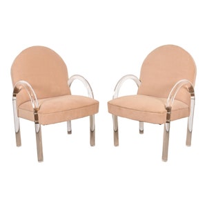May include: Two armchairs with a peach-colored, rounded back and seat. The arms and legs are clear acrylic, creating a modern, airy aesthetic. The chairs are set against a white background.