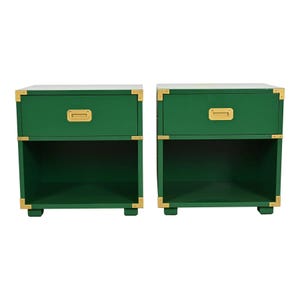 May include: Two green nightstands with brass-colored hardware. Each nightstand has a drawer with a brass pull and an open shelf below. The corners are reinforced with brass accents, and they sit on short, square feet.