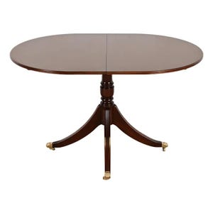 May include: Oval-shaped wooden dining table with a dark brown finish. The table has a pedestal base with three curved legs and gold-colored accents on the feet. The tabletop is approximately 48 inches long.