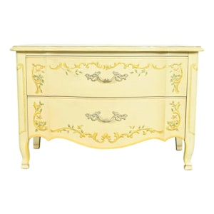 May include: Cream-colored two-drawer dresser with floral accents. The dresser features ornate gold and green painted floral designs and silver-toned drawer pulls. The curved legs and edges add to the vintage aesthetic.