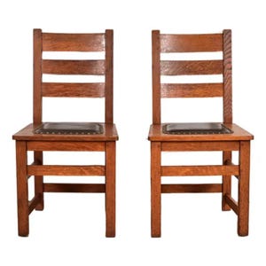 May include: Two antique wooden chairs with a warm, reddish-brown finish. Each chair features a rectangular seat with a dark, possibly leather, cushion and a ladder-back design with horizontal slats. The chairs are sturdy and have a classic, vintage aesthetic.