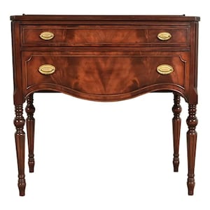 May include: A mahogany-colored antique side table with two drawers and brass hardware. The table has a curved apron and four turned legs. The drawers have oval brass pulls. The table is a classic piece of furniture.