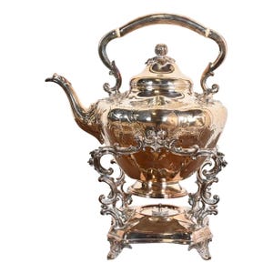 May include: A silver teapot with an ornate design and a stand. The teapot has a round body with a spout and a handle. The stand has a square base with four legs.