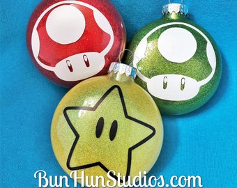 Könnte beinhalten: Drei Weihnachtsbaumschmuck mit Figuren aus dem Videospiel Super Mario Bros. Der Schmuck ist rot, grün und gelb mit Glitzer. Der rote Schmuck ist ein Pilz, der grüne Schmuck ist ein Pilz und der gelbe Schmuck ist ein Stern.