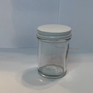 Puede incluir: Un tarro de cristal transparente con tapa metálica blanca. El tarro es cilíndrico con base ligeramente redondeada. La tapa es plana y circular, con borde plateado. El tarro está vacío y sobre una superficie blanca.