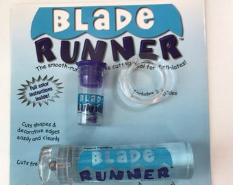 May include: A Blade Runner cutting tool package. The package includes a clear plastic cutting tool with the words "Blade Runner" in blue and white, a small purple container, and a clear plastic ring. The package is blue and white.