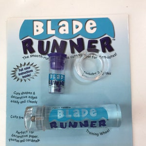 May include: A Blade Runner cutting tool package. The package includes a clear plastic cutting tool with the words "Blade Runner" in blue and white, a small purple container, and a clear plastic ring. The package is blue and white.