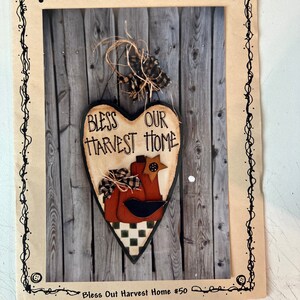 May include: A rustic heart-shaped wooden sign with the text "Bless Our Harvest Home." The sign features a pumpkin and bird design, with a black and white checkered bottom. The sign is attached to a wooden fence background.