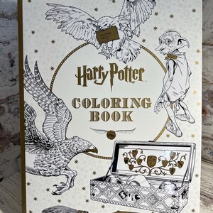 May include: A Harry Potter coloring book with a white cover featuring black and gold illustrations of an owl, a house-elf, a hippogriff, a broomstick, and an open trunk. The text Harry Potter Coloring Book is printed in the center in gold.