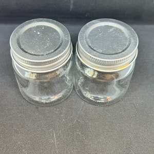 Puede incluir: Dos tarros de vidrio transparente con tapas de metal plateado. Los tarros son redondos, con boca ancha y forma corta y rechoncha. Las tapas tienen una parte superior plana y un borde roscado para atornillar al tarro. Los tarros están sobre una superficie negra.