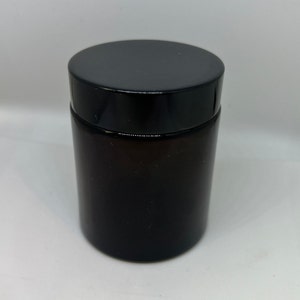 Puede incluir: Un tarro de vidrio marrón oscuro con tapa negra. El tarro es cilíndrico y opaco, con un acabado liso y brillante. La tapa es plana y también negra. El tarro está sobre una superficie blanca.