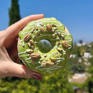 May include: A close-up of a green glazed donut topped with nuts and white icing drizzle. The donut is held in a hand against a backdrop of green foliage and a blue sky. A sweet treat.