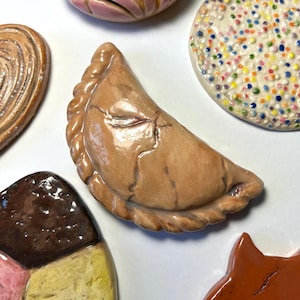 May include: A collection of ceramic food ornaments including a tan empanada with a crimped edge, a pink concha, a round cookie with multicolored sprinkles, a brown spiral pastry, a multi-colored ice cream scoop, and a smooth orange-brown piece.