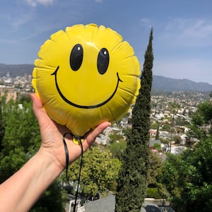 May include: A round, bright yellow foil balloon featuring a classic black smiley face graphic with two oval eyes and a wide, curved mouth. The balloon has a crinkled edge and is attached to a thin black ribbon hanging from the bottom center.
