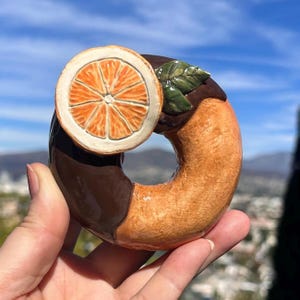 May include: A ceramic donut with a chocolate glaze on one half and a light orange glaze on the other. It is topped with a ceramic orange slice and a green leaf. The donut is held up against a blue sky.