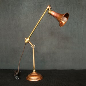 May include: An adjustable desk lamp with a copper shade and base, and brass-colored arms. The lamp has a vintage industrial design, with a visible lightbulb and a brown power cord. The lamp is on a black surface.