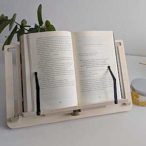 Può includere: Un leggio in legno chiaro che tiene un libro aperto. Il leggio ha un design semplice con bracci regolabili e una base. Il libro è aperto su una pagina con testo. Una pianta e un barattolo sono sullo sfondo.