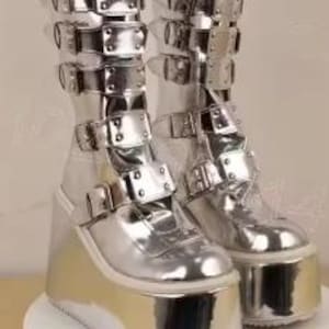 May include: A pair of knee-high boots with a metallic silver finish, featuring a thick platform wedge sole and multiple horizontal straps across the front. Each strap is secured with a polished silver-tone metal buckle, extending from the toe to the calf.