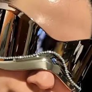 May include: Metallic silver sunglasses with a curved, wraparound design. The sunglasses feature a decorative line of small, clear rhinestones along the top edge. The lenses reflect the surrounding environment.