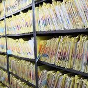 May include: Rows of file folders with yellow tabs fill shelves in a storage room. The folders are neatly organized, creating a grid-like pattern. The room has a red floor and the shelves are black.