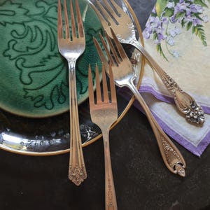 Può includere: Quattro forchette d'argento con manici decorati sono disposte su un piatto verde e dorato e un tovagliolo floreale bianco e viola. Le forchette sono tutte di stili diversi e hanno un aspetto vintage.