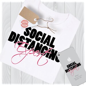 Könnte beinhalten: Weißes T-Shirt mit den Worten "SOCIAL DISTANCING Expert" in schwarzer und rosa Schrift. Ein braunes Etikett mit "APPROVED TESTED" ist angebracht. Ein silbernes iPhone zeigt denselben Text. Das Shirt liegt auf einer hellen Holzoberfläche.