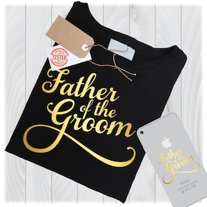 Puede incluir: Camiseta negra con texto dorado que dice "Father of the Groom". Un diseño a juego está en un smartphone. Una etiqueta con las palabras "Approved Tested" está adherida a la camiseta.
