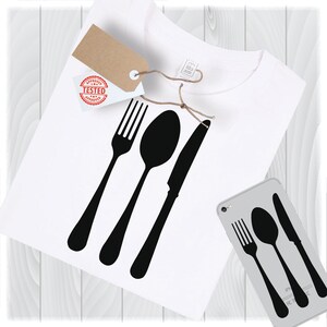 May include: A white t-shirt with a black silhouette of a fork, spoon, and knife. The design is centered on the shirt. The shirt is on a white wooden surface.