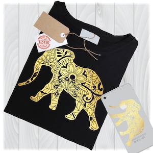 May include: A black t-shirt with a gold elephant design. The elephant is decorated with intricate floral patterns. The shirt is folded and lying on a wooden surface.