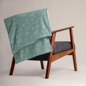 Könnte beinhalten: Eine tealblaue Wolldecke mit einem weißen Muster aus verschiedenen magischen Gegenständen, darunter Schwerter, Zauberstäbe und Sterne. Die Decke liegt über einem Holzsessel.
