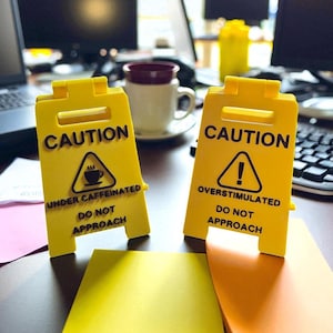 Puede incluir: Dos señales de precaución amarillas con texto y símbolos negros. Una señal dice "CAUTION UNDER CAFFEINATED DO NOT APPROACH" con un símbolo de taza de café. La otra dice "CAUTION OVERSTIMULATED DO NOT APPROACH" con un signo de exclamación.