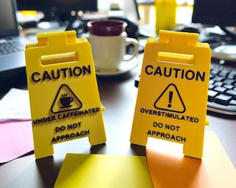 Puede incluir: Dos señales de precaución amarillas con texto y símbolos negros. Una señal dice "CAUTION UNDER CAFFEINATED DO NOT APPROACH" con un símbolo de taza de café. La otra dice "CAUTION OVERSTIMULATED DO NOT APPROACH" con un signo de exclamación.