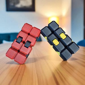 May include: Two fidget toys, one red and one black, are displayed on a wooden surface. The red toy has black connectors, while the black toy features yellow accents. The toys are designed with a modular, interlocking structure.