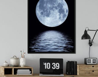 Puede incluir: Una impresión enmarcada que representa una luna llena reflejada en agua ondulante sobre un fondo negro. La luna es de color azul pálido y el reflejo del agua refleja su forma. La impresión se muestra encima de un escritorio de madera con un portátil que muestra la hora.