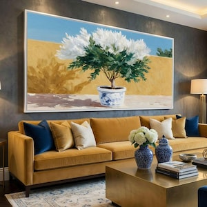 May include: A large framed painting depicts a tree with white flowers and green leaves in a blue and white pot. The painting hangs above a mustard-colored sofa with blue and cream pillows. A gold coffee table and blue and white vases complete the scene.