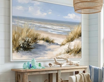 Può includere: Grande dipinto incorniciato di una scena di spiaggia con bordo bianco. L'opera raffigura un sentiero sabbioso che conduce al mare, con erba alta e cielo blu. Il dipinto è sopra una consolle di legno.