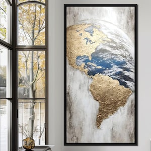 May include: Framed artwork showing a textured gold and blue Earth globe. The piece is set against a neutral backdrop and framed in black. It's positioned near a window with a view of trees.
