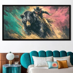 May include: A framed canvas print depicts a cowboy riding a large black bear. The artwork features a dynamic, painterly style with a color palette of teal, gold, and pink. The cowboy wears a hat and scarf, holding reins.