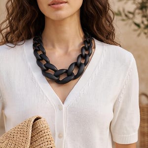 Puede incluir: Un cárdigan blanco de manga corta con cuello en V y botones, un collar de eslabones negros mate gruesos y un bolso de mano tipo sobre de paja tejida color canela. El texto Urban Sense con una línea y símbolo aparece abajo a la derecha.