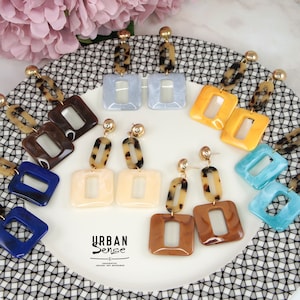 Puede incluir: Surtido de pendientes cuadrados de colores con una variedad de patrones y diseños. Los pendientes se muestran en un plato blanco con un patrón de cuadros blanco y negro. Los pendientes están hechos de plástico y tienen ganchos de metal dorado.