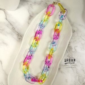 Puede incluir: Un collar de cadena de plástico de colores arcoíris. La cadena está hecha de eslabones grandes de forma ovalada. El collar está sobre una superficie de mármol blanco.