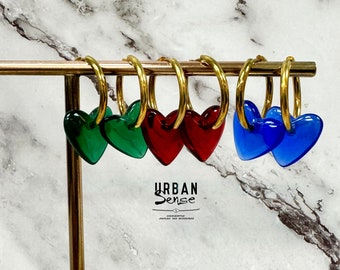 Puede incluir: Aretes de aro dorados con dijes en forma de corazón en rojo, verde y azul. Los aretes se muestran en un soporte dorado. El texto "URBAN Sense" es visible debajo de los aretes.
