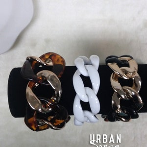 May include: Three chunky chain bracelets in different colors. The bracelets are made of plastic and have a gold-tone metal accent. The bracelets are displayed on a black surface. The text "URBAN Sense" is visible in the bottom center of the image.