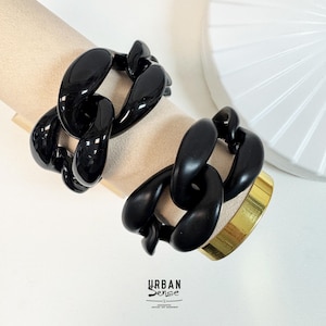May include: Two black chain link bracelets. One bracelet is resting on a beige surface. The other bracelet is paired with a gold-colored cuff. The bracelets are glossy and have a bold, chunky design. The logo "URBAN Sense" is visible.