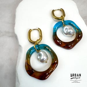 Puede incluir: Un par de pendientes con aros ovalados dorados unidos a dijes circulares irregulares en un degradado de turquesa y marrón translúcido. Una perla barroca blanca cuelga del centro de cada aro. El texto dice URBAN SENSE.