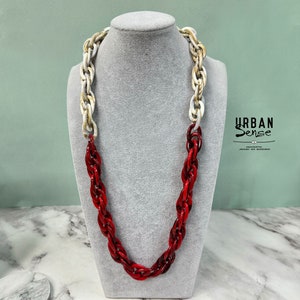 Pode incluir: Um colar de corrente grosso vermelho e branco. O colar é feito de plástico e tem um design de corda. O colar é exibido em um manequim branco.