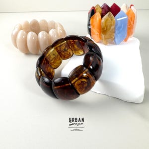 May include: Three stretch bracelets made of polished resin segments. One bracelet is solid cream, one is dark brown with a marbled pattern, and one features a multi-colored array of orange, red, yellow, and blue segments. Text reads URBAN SENSE.